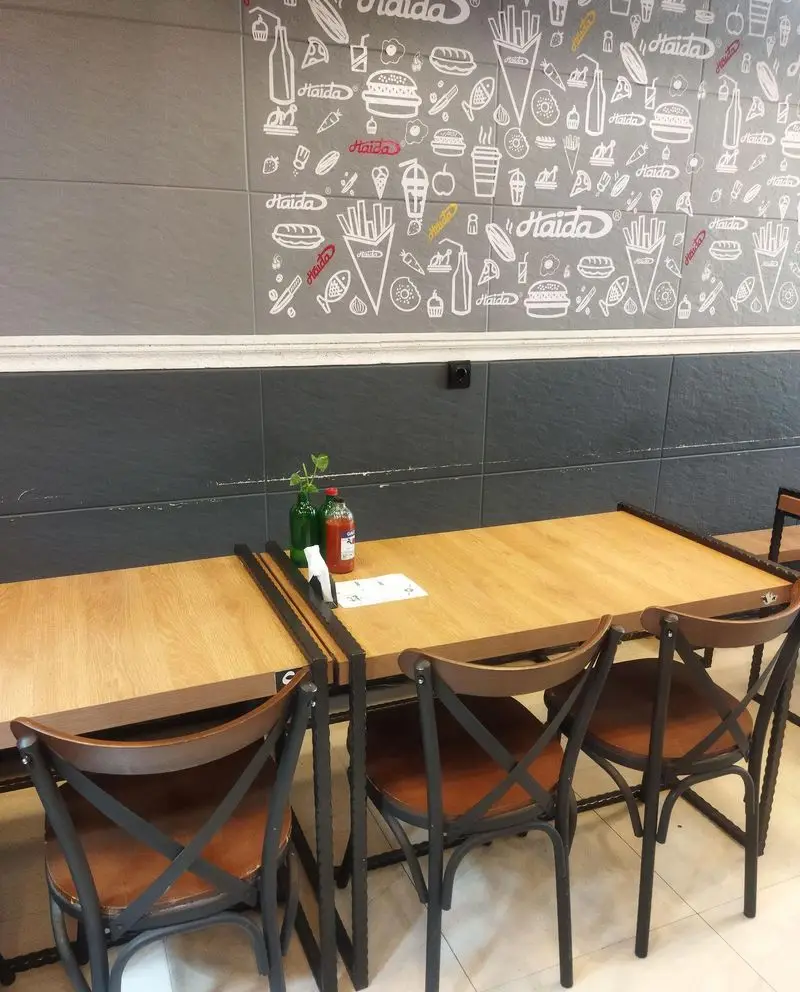 دکوراسیون داخلی فست فود هایدا پیروزی با میز و صندلی‌های چوبی و فلزی، دیوار نقاشی شده با طرح‌های فست فود و لوگوی هایدا.