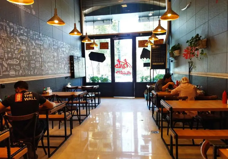 فضای داخلی دنج و مدرن فست فود هایدا پیروزی با مشتریان در حال صرف غذا، میز و صندلی‌های چوبی، و دکوراسیون دیواری طرح‌دار.