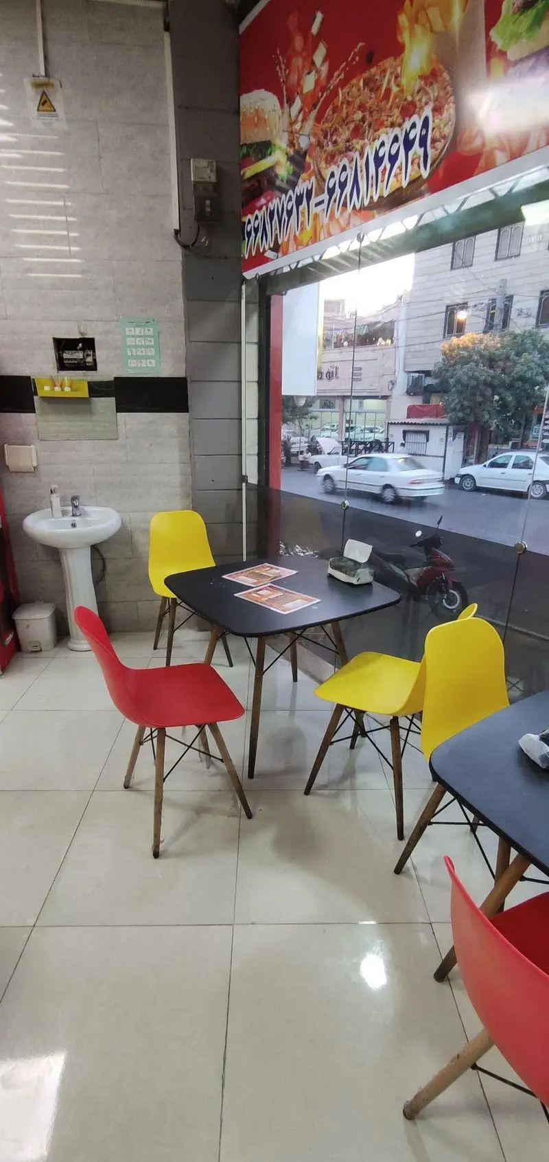 فضای داخلی فست فود مزه: میز و صندلی‌های مدرن (قرمز و زرد)، سینک، پنجره رو به خیابان. بنر تبلیغاتی با عکس برگر و پیتزا.