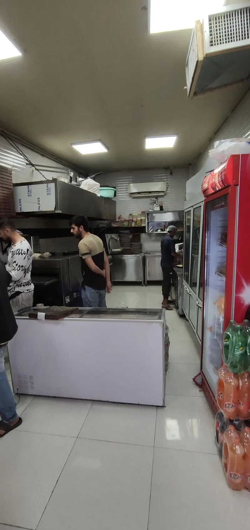 نمای داخلی آشپزخانه فست فود مزه با کارکنان مشغول تهیه غذا، تجهیزات استیل، فریزر و یخچال نوشیدنی. محیطی تمیز و فعال.