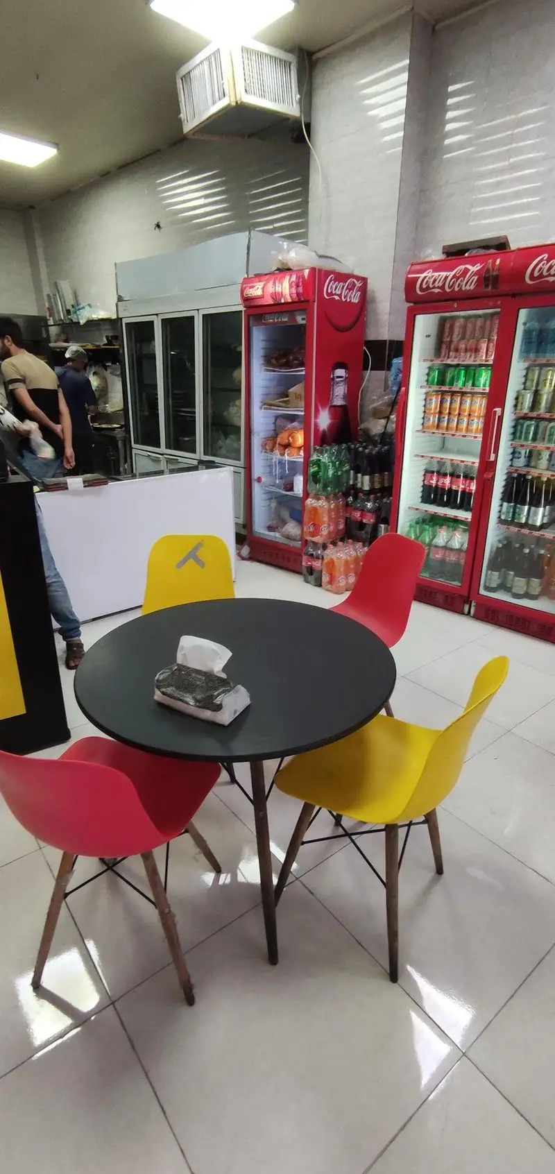 فضای داخلی فست فود مزه با میز و صندلی‌های رنگی، یخچال‌های نوشیدنی کوکاکولا و پرسنل در حال کار در آشپزخانه.