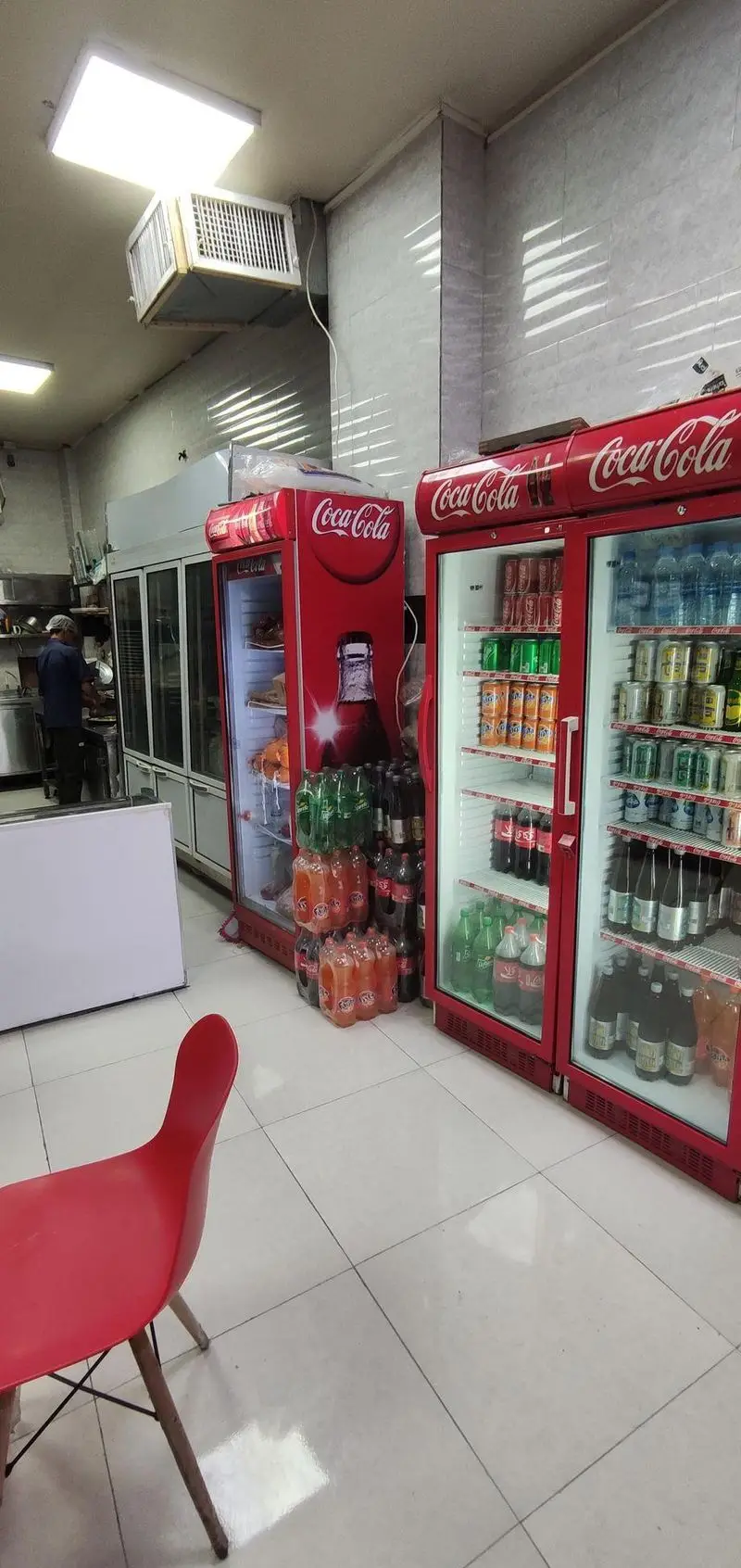 فضای داخلی فست فود مزه با یخچال‌های قرمز کوکا کولا پر از نوشیدنی، قفسه‌های غذاهای آماده و آشپزی در حال کار.
