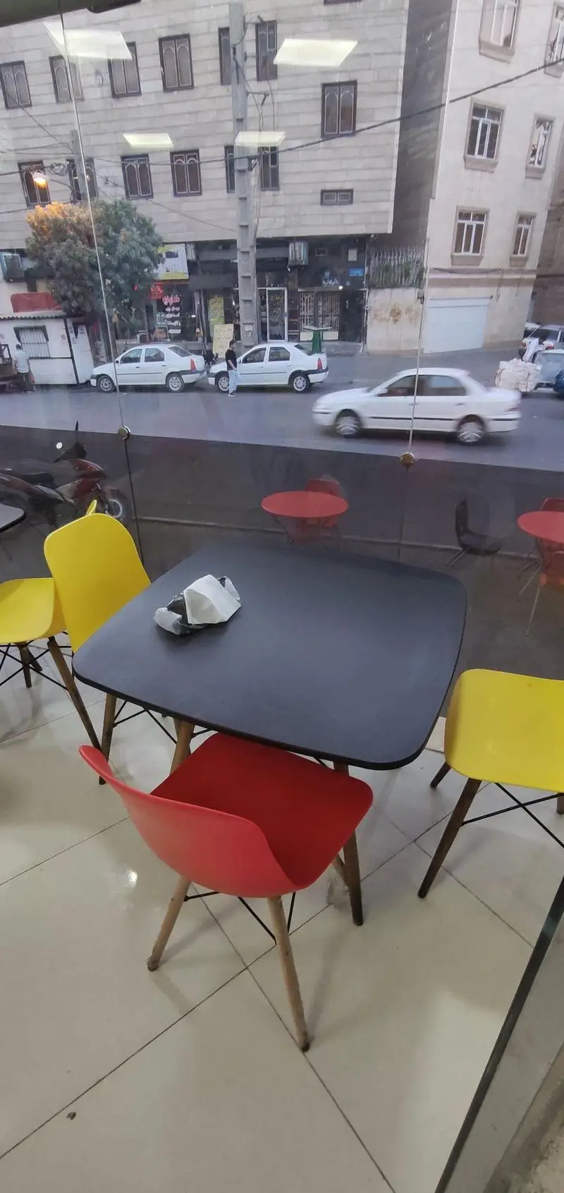 نمای داخلی فست فود مزه با میز مشکی و صندلی‌های رنگی زرد و قرمز، همراه با چشم‌اندازی از خیابان شهری و ماشین‌ها از پشت پنجره.