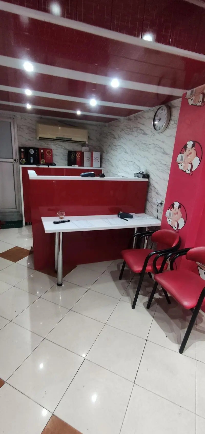 نمای داخلی کترینگ ارغوان با دکوراسیون مدرن قرمز و سفید، میز پذیرش، صندلی‌های مشتری و تصاویر جذاب سرآشپز برای ثبت سفارش غذا.