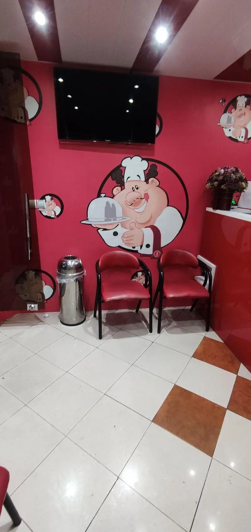 تصویری از فضای داخلی کترینگ ارغوان، با دکوراسیون شاد و قرمز، طرح‌های جذاب آشپز، تلویزیون، صندلی‌های راحت و کفپوش زیبا. محیطی دلنشین برای پذیرایی.