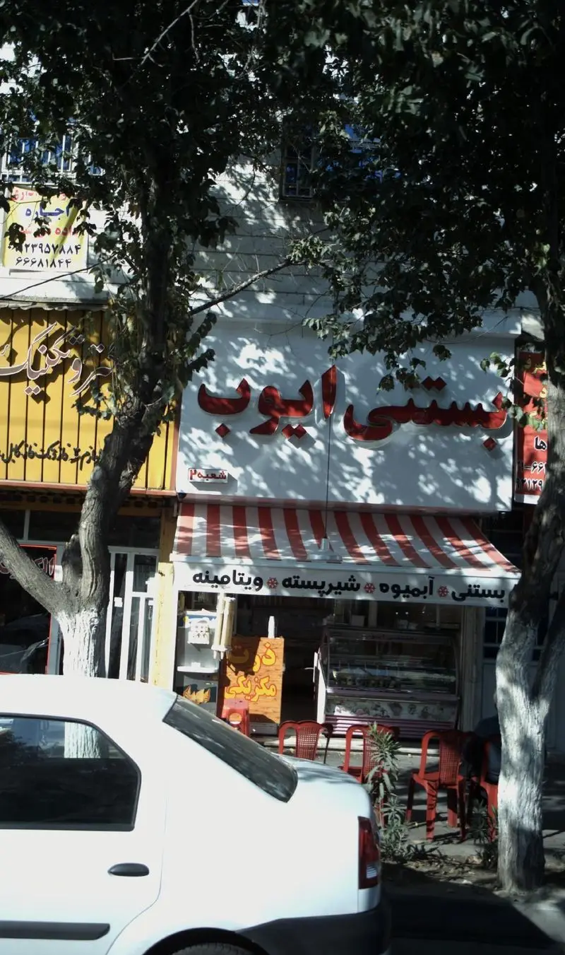 مغازه بستنی ایوب با تابلوی قرمز و سایبان راه راه، فروش بستنی، آبمیوه، شیر پسته و ویتامینه، با خودرویی در جلو و درختان سبز.