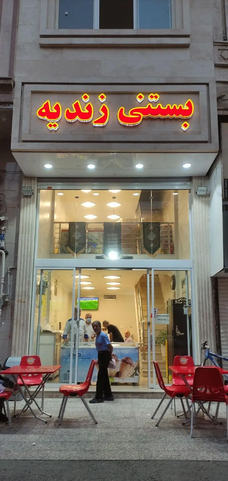 تصویر بستنی فروشی زندیه با تابلوی جذاب و روشن، نمایشگر بستنی‌های متنوع و مشتریان و میز و صندلی در فضای بیرونی.