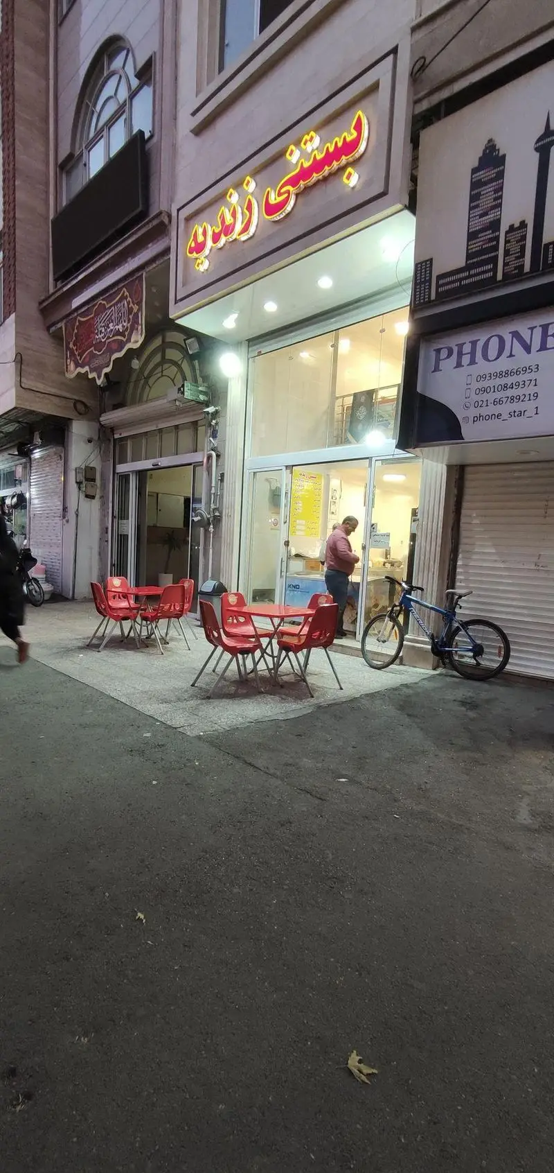 نمای بیرونی بستنی فروشی زندیه با تابلوی درخشان، ورودی شیشه‌ای، میز و صندلی‌های قرمز در پیاده‌رو برای مشتریان و دوچرخه‌ای در کنار مغازه.