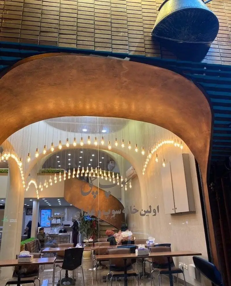 نمای داخلی رستوران مدرن سوپ چی با دکوراسیون گرم و نورپردازی زیبا، میز و صندلی‌های چوبی و مشتریان در حال صرف غذا در اولین خانه سوپ.