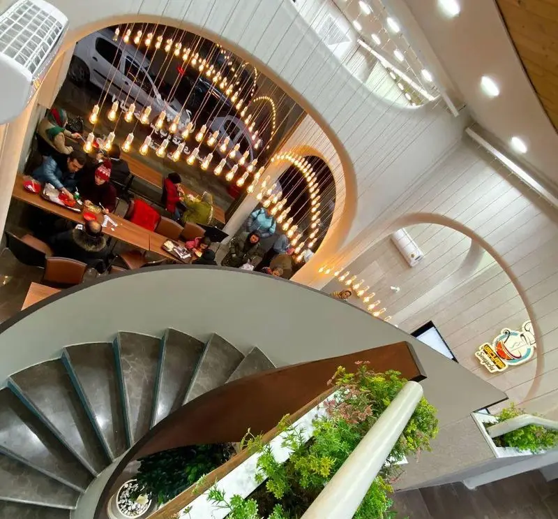 نمای چشم‌نواز از رستوران سوپ چی با پله مارپیچ سنگی، نورپردازی هنری با لامپ‌های آویز و فضای پر جنب و جوش مشتریان.