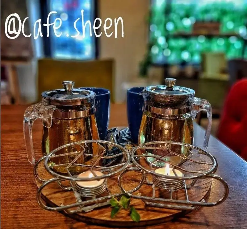 دو قوری چای دمنوش با گرم نگهدارنده شمعی و فنجان های آبی در کافه شین، مناسب برای تجربه نوشیدنی گرم و دلپذیر.