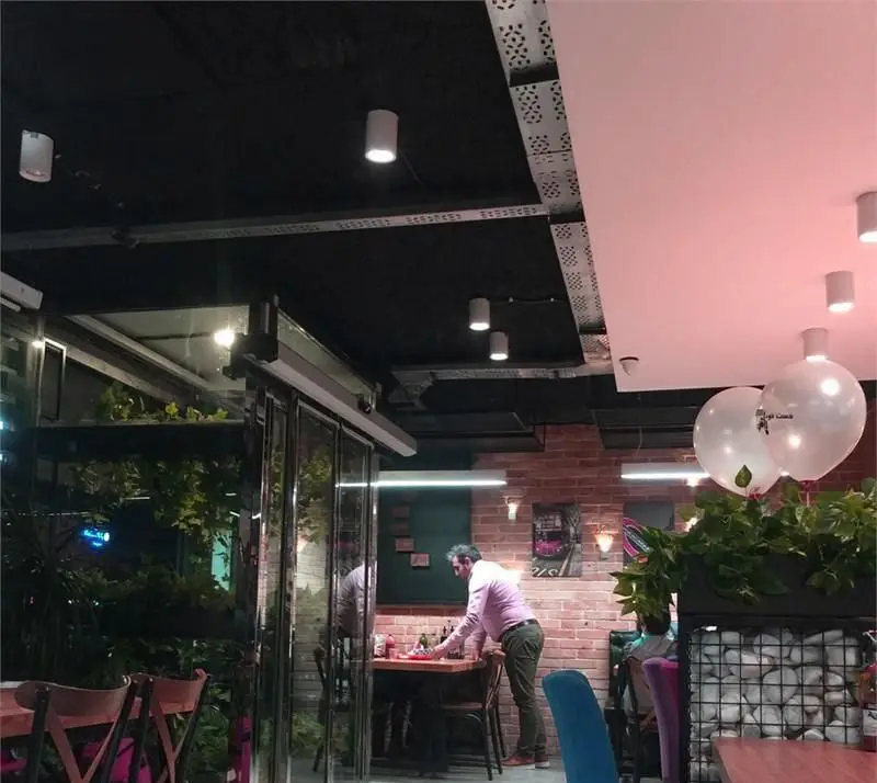 فضای داخلی و دکوراسیون مدرن فست فود بامبی فود، شامل سقف‌های مشکی و صورتی، دیوارهای آجری، میز و صندلی شیک، گیاهان سبز و بالکن شیشه‌ای زیبا.