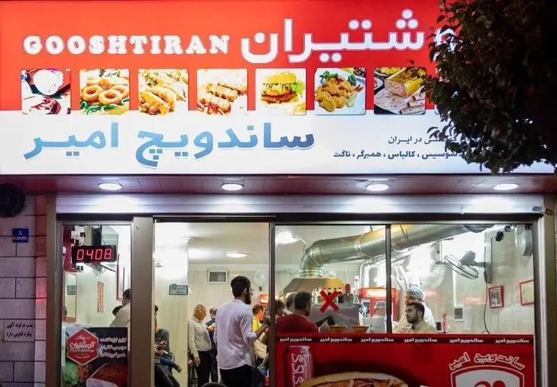 نمایی از ساندویچ امیر، فست فود محبوب با محصولات گوشتیران. منو شامل سوسیس، کالباس، همبرگر و ناگت. شلوغی داخل مغازه.