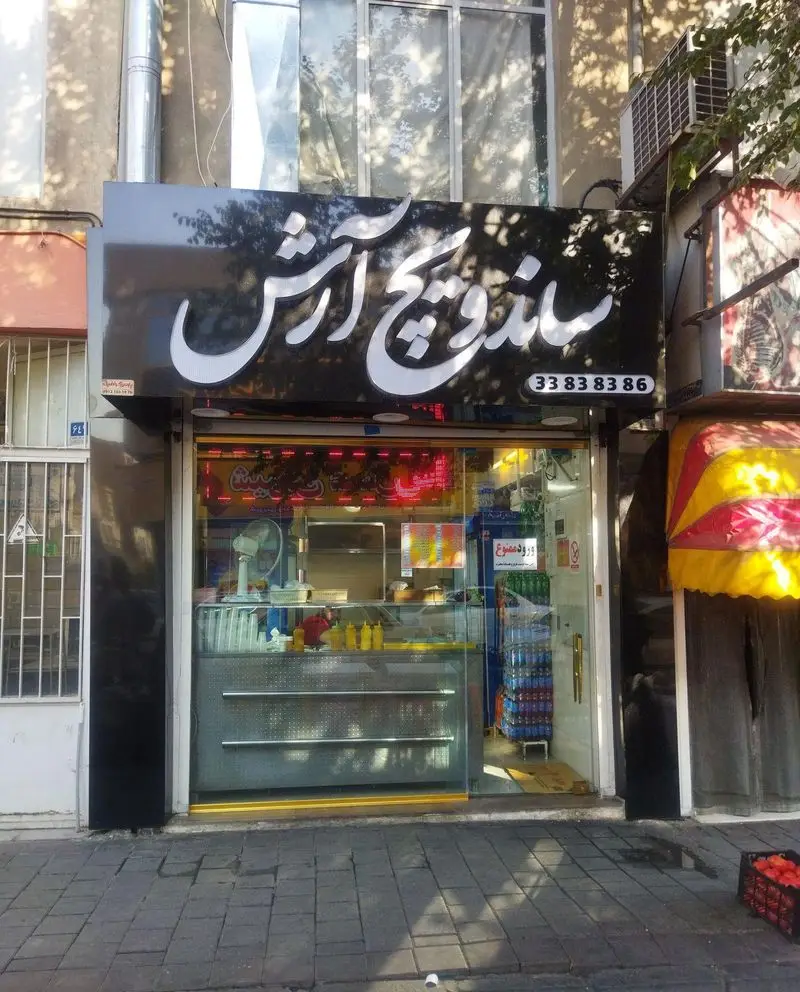 نمای بیرونی مغازه ساندویچ و فلافل آرش با تابلو مشکی و حروف سفید روشن، شامل شماره تماس 33838386 و ویترین آماده.