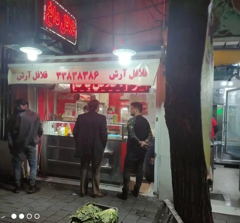 نمای بیرونی فلافل فروشی آرش در شب با نورپردازی گرم، تابلوی "فلافل داغ" و مشتریان در حال خرید غذای خیابانی. شماره تلفن: ۳۳۸۳۸۳۸۶.