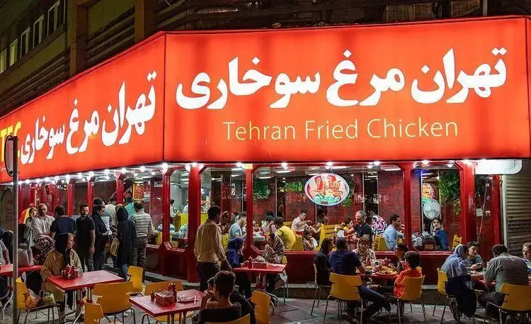 تصویر یک شعبه شلوغ و پرنور از رستوران مرغ سوخاری تهران با تابلوی قرمز رنگ و مشتریان در حال صرف غذا در شب.