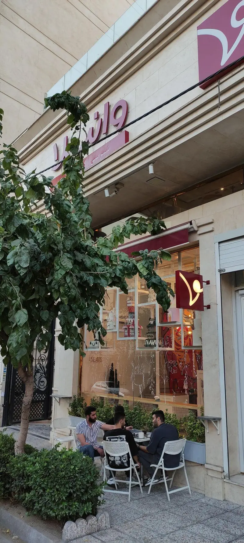 کافه قنادی وانیلا: نمای خارجی یک شیرینی‌فروشی و کافه مدرن در فضای باز، مردانی در حال گپ‌وگفت و لذت از نوشیدنی، ویترین جذاب کیک.