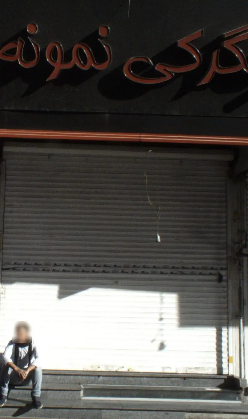 تابلوی مغازه جگرکی نمونه با حروف برجسته قرمز، درب کرکره‌ای بسته و مردی روی پله‌های ورودی. نمای بیرونی جگرکی.