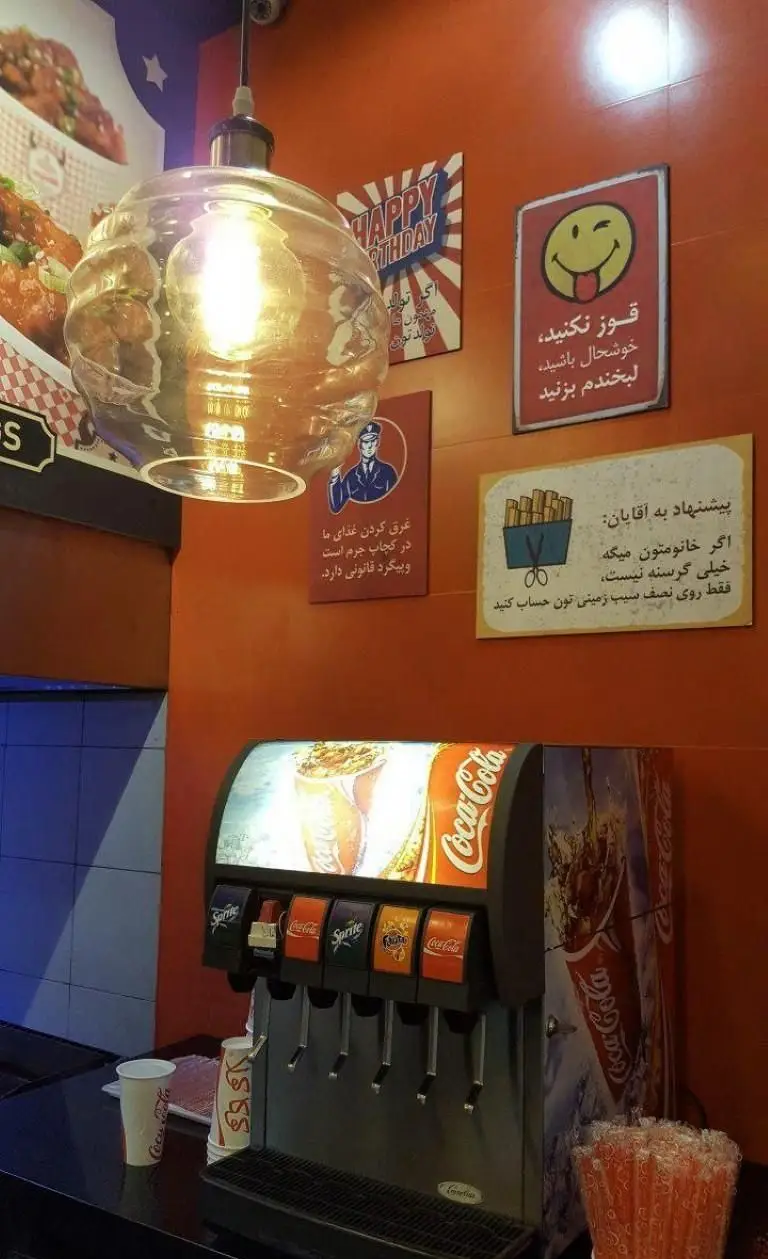 تصویر داخلی رستوران فست فود گامبی، با دستگاه نوشابه کوکاکولا، اسپرایت و فانتا و تابلوهای دیواری رنگارنگ و منوی مرغ سوخاری.