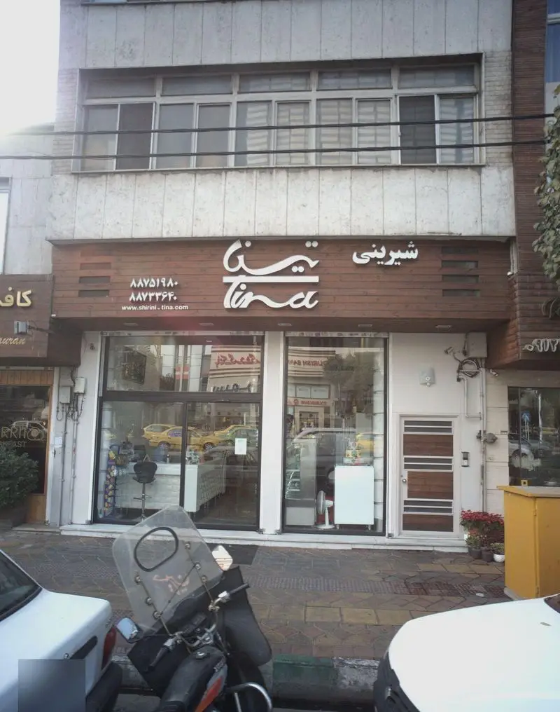 تصویر نمای جلوی شیرینی فروشی و قنادی تینا در شهر تهران با ویترین بزرگ شیشه‌ای و اطلاعات تماس روی تابلوی چوبی.