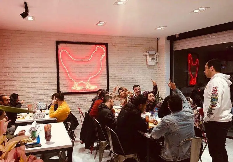 مشتریان در فست فود چیکن باکس مشغول صرف غذا و گفتگو، با تابلوی نئون مرغ در پس زمینه آجری سفید.