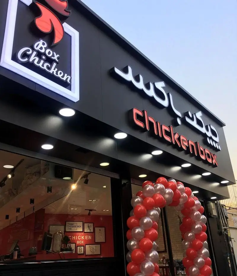 نمای بیرونی فست فود Chicken Box با سردر مشکی، تابلوی روشن چیکن باکس فارسی و انگلیسی و بادکنک آرایی افتتاحیه.