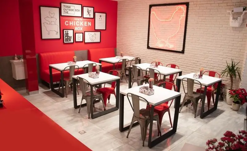 فضای داخلی دنج و مدرن فست فود چیکن باکس با دیوارهای قرمز، میز و صندلی‌های شیک و نئون مرغ جذاب برای مشتریان.