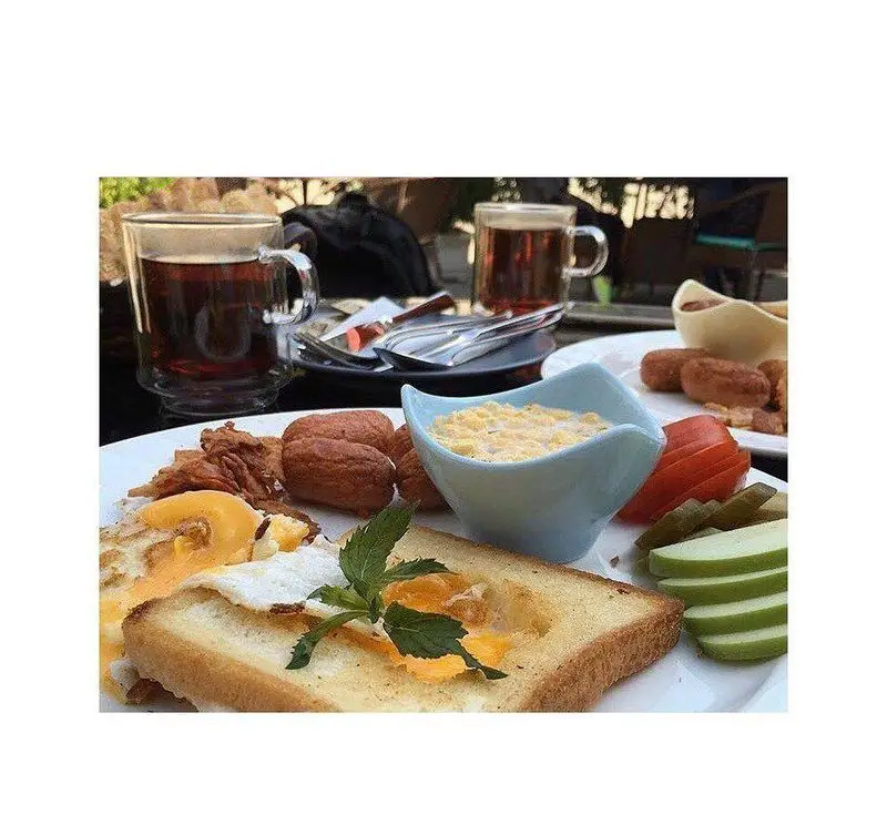 صبحانه مفصل و خوشمزه کافه رستوران مازو شامل تخم مرغ، نان تست، سوسیس، سیب، خیارشور و چای در فضای باز.