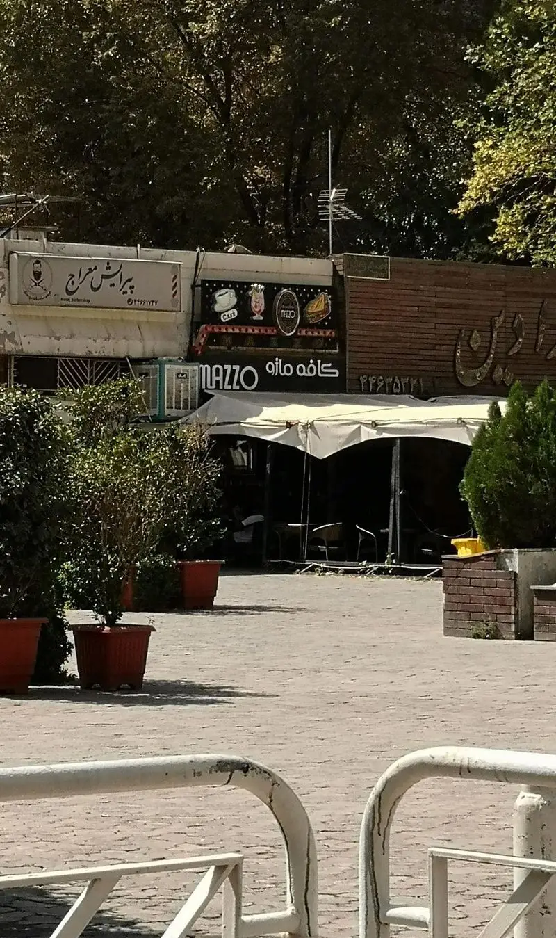 نمای واضح از ورودی کافه رستوران مازو همراه با چترهای فضای باز، گیاهان تزئینی و تابلوی کافه مازو و پیرایش معراج در یک روز آفتابی.