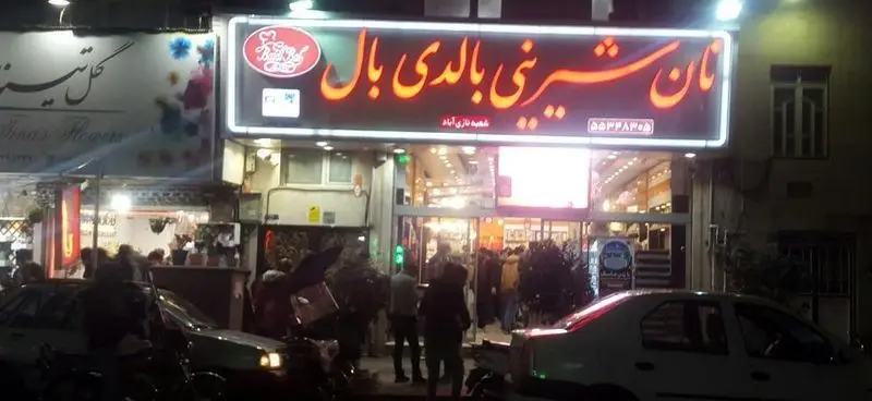 نمای بیرونی شیرینی بالدی بال شعبه نازی‌آباد در شب؛ یک نان و شیرینی فروشی روشن با تابلو نئون و مشتریان در حال خرید.