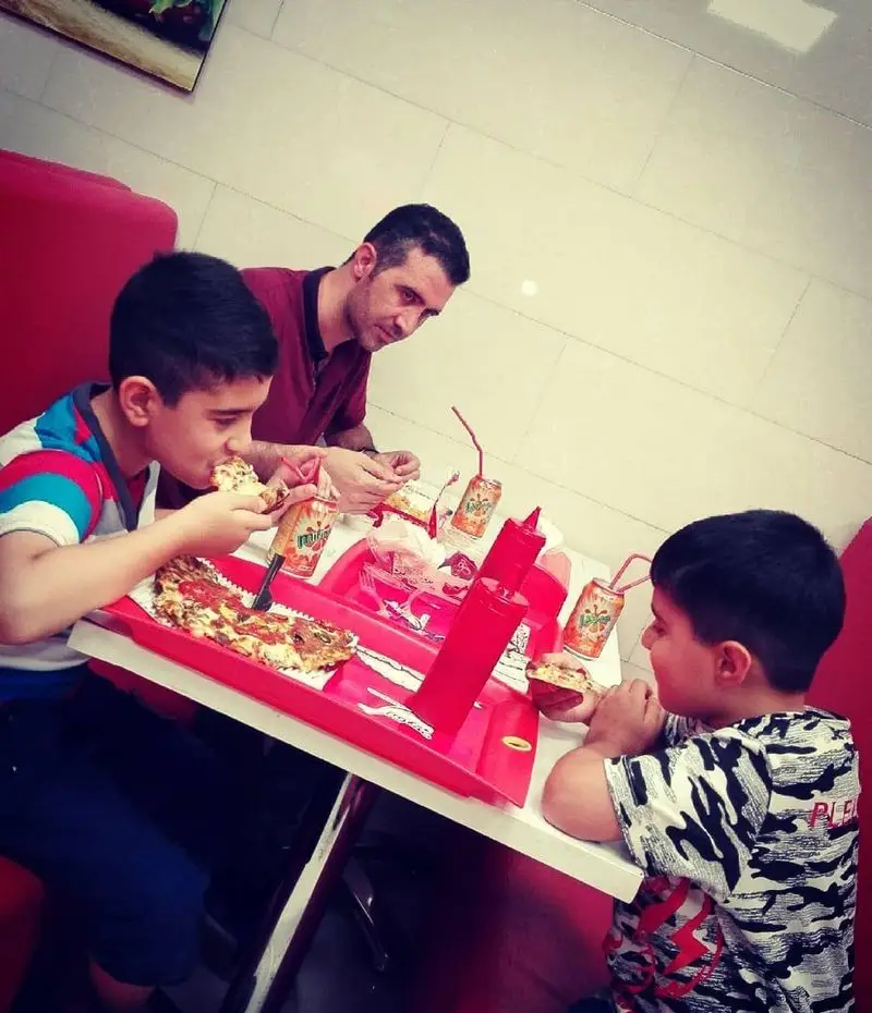 تصویری از خانواده‌ای شاد در فست فود کنتاویچ، پدر و دو فرزندشان در حال لذت بردن از پیتزاهای خوشمزه و نوشیدنی‌های خنک در محیطی دلپذیر.