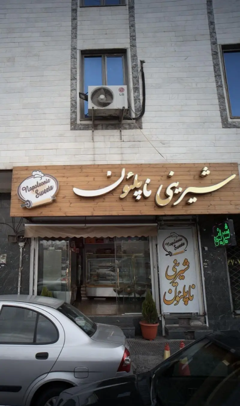 نمای بیرونی فروشگاه شیرینی ناپلئون (Napoleonic Sweets) با تابلوی چوبی برجسته و نورانی. ورودی مغازه با ویترین شفاف و خودروها.