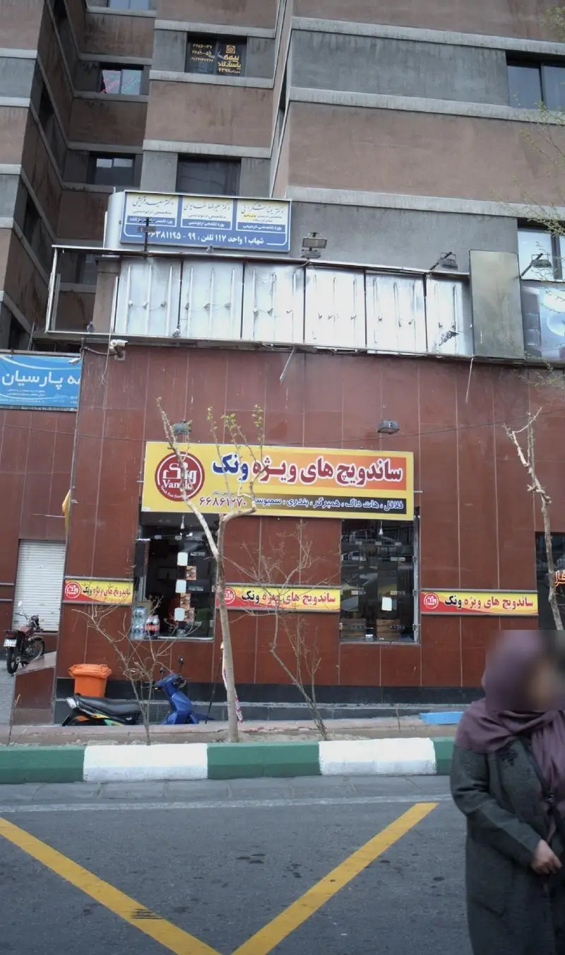 تصویر مغازه ساندویچ های ویژه ونک با تابلوی زرد روشن در خیابان، عرضه کننده انواع ساندویچ، فلافل، هات داگ، همبرگر و سمبوسه.