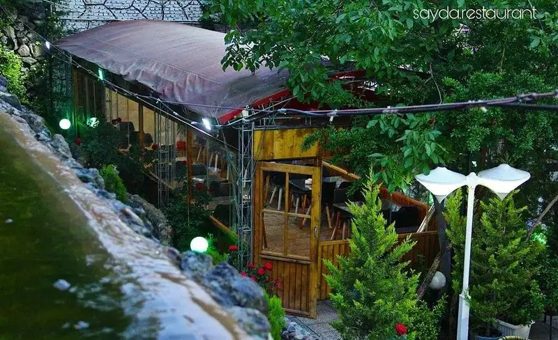 تصویری از فضای دلپذیر و سبز باغ رستوران سایدا شهران با محوطه غذاخوری روباز، آبنما، درختان و گل‌های زیبا برای تجربه‌ای خاطره‌انگیز.