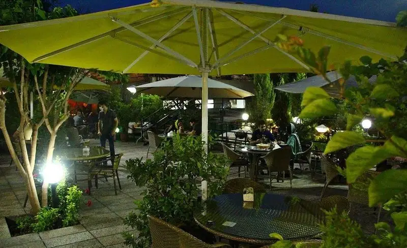 فضای دلنشین باغ رستوران سایدا شهران در شب، با چترهای زرد و میزهای آماده برای پذیرایی از مشتریان در محیطی سرسبز و آرام.