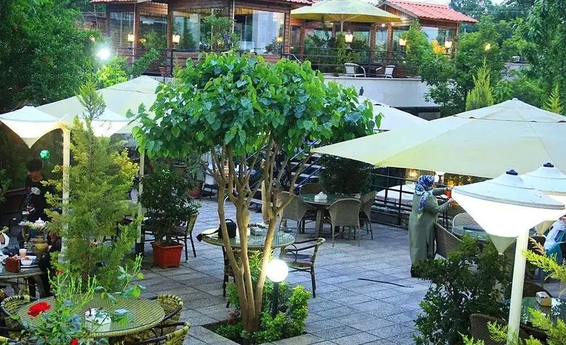 باغ رستوران سایدا شهران: فضای باز و سرسبز با میز و صندلی چیده شده زیر چترهای آفتابگیر، محیطی دلنشین برای دورهمی و لذت بردن از غذا.