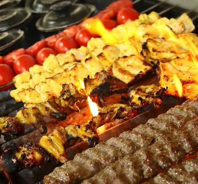 منظره جذاب از کباب کوبیده و جوجه کباب زعفرانی خوشمزه به همراه گوجه فرنگی کبابی در حال پخت روی آتش زنده در باغ رستوران سایدا شهران.