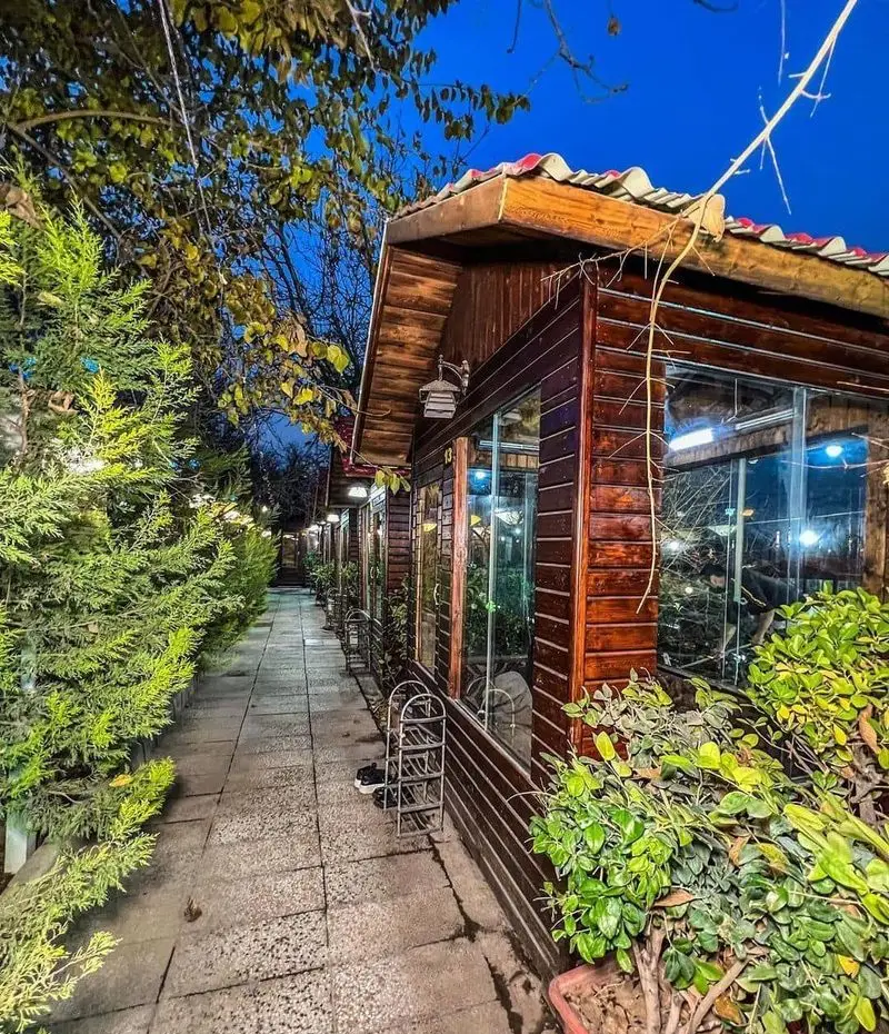 باغ رستوران سایدا شهران: تصویری جذاب از مسیر سنگی و کلبه‌های چوبی زیبا در فضای سبز شب، با نورپردازی دلنشین برای تجربه‌ای آرام.