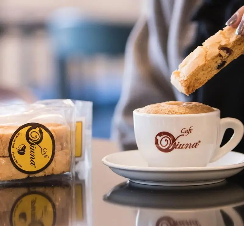فنجان قهوه داغ کافه ویونا، همراه با بیسکویت ویونا که در آن دیپ می‌شود. بسته بندی محصولات کافه ویونا نیز در تصویر است.