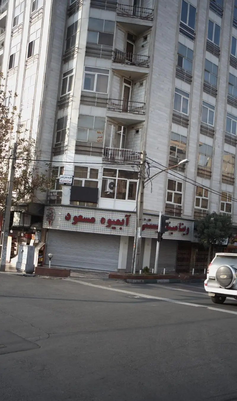 نمای خارجی ویتامینه، بستنی و آبمیوه مسعود در یک ساختمان چندطبقه با کرکره‌های بسته و خیابانی آرام، نشانگر یک روز تعطیل.
