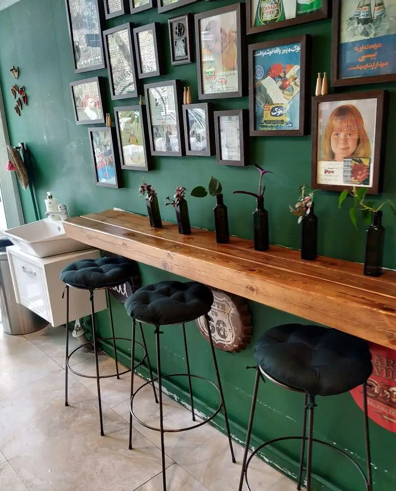 فضای داخلی دلنشین ساندویچی باماهاس با دیوار سبز زمردی، قاب‌های هنری و پوستر‌های قدیمی، میز چوبی بلند و صندلی‌های بار مدرن و راحت، محیطی عالی برای مشتریان.