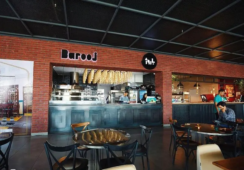 تصویری از فضای داخلی بزرگ و مدرن فست فود باروژ، شامل پیشخوان آماده‌سازی غذا، پرسنل فعال و مشتریان در حال لذت بردن از محیط و غذا. طراحی با دیوارهای آجری.