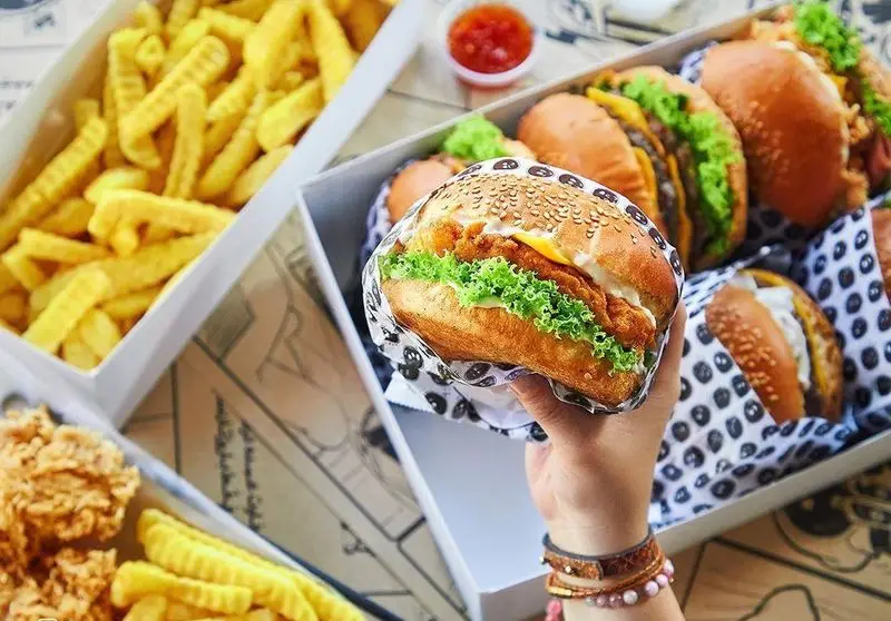 دست در حال گرفتن همبرگر سوخاری از پک‌های فست فود باروژ، همراه با باکس سیب زمینی سرخ کرده و مرغ سوخاری. لذت طعم واقعی.