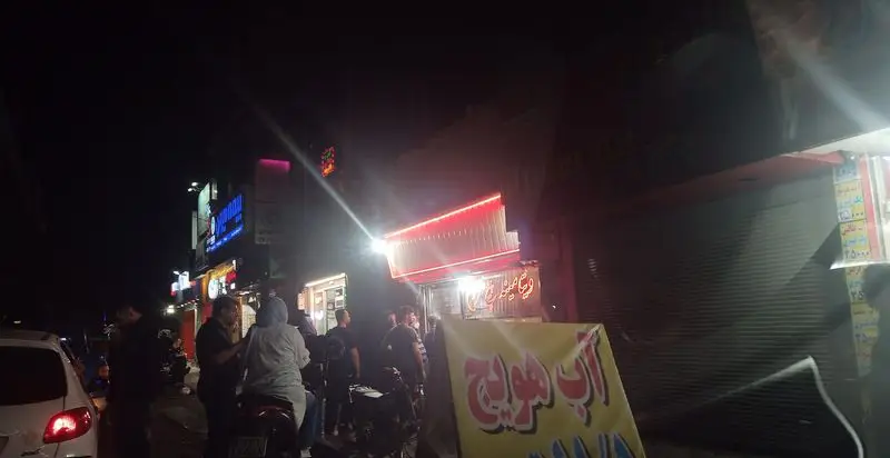 نمای شبانه و پر جنب و جوش از مغازه بستنی و معجون پیچ شمرون در خیابان شلوغ با مشتریان و نورپردازی خیره‌کننده