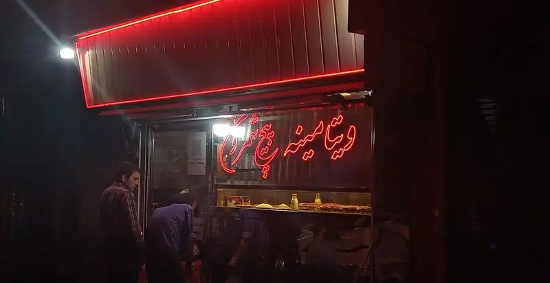 تصویر شب از نمای بیرونی مغازه بستنی و معجون پیچ شمرون با تابلوی نئون قرمز روشن، مشتریان و ویترین محصولات دسر و خوراکی.