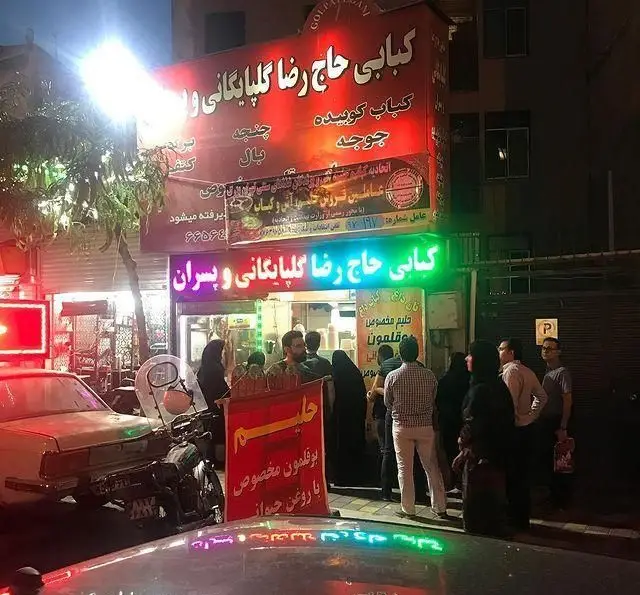کبابی حاج رضا گلپایگانی و پسران در شب با تابلوهای نئون و صف مشتریان که منتظر کباب و حلیم هستند.