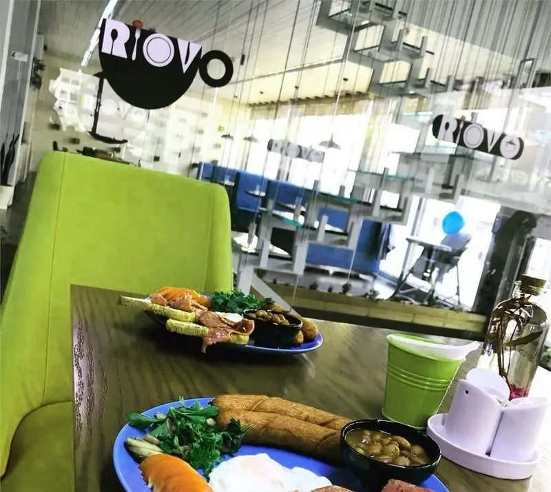 نمایی از فضای داخلی کافی شاپ ریو وو با میز صبحانه کامل و لذیذ. صندلی‌های سبز رنگ و شیشه‌های شفاف، فضایی دلنشین و مدرن را به ارمغان آورده‌اند.