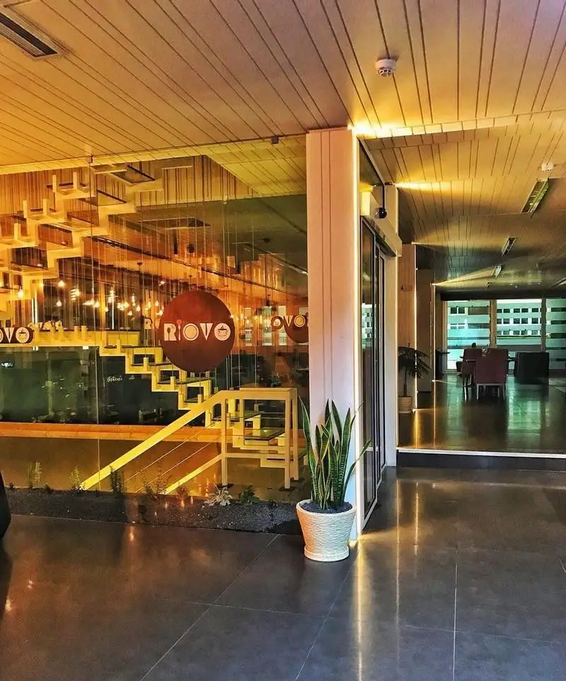 فضای داخلی دلنشین و مدرن کافی شاپ ریو وو با سقف چوبی، نورپردازی گرم، دکوراسیون شیک، پله‌های چوبی تزئینی و لوگوی RIOVO روی شیشه.