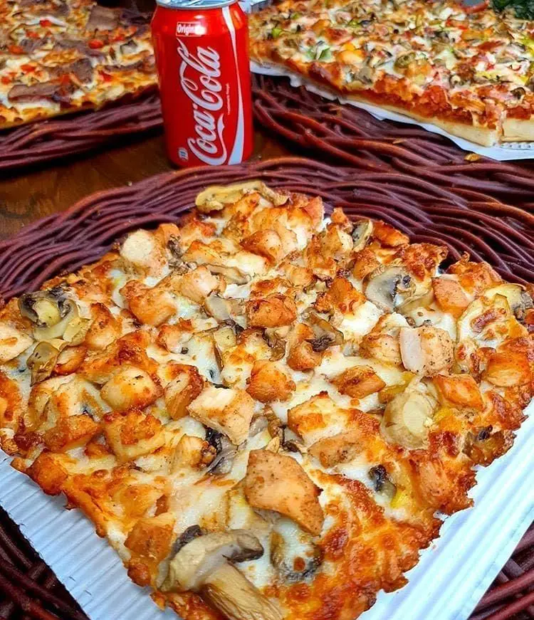 پیتزا مرغ و قارچ خوشمزه از پیتزا کوک، با قوطی کوکاکولا در کنار و پیتزاهای متنوع دیگر در پس‌زمینه، بهترین انتخاب برای غذای امروز شما.