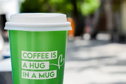فنجان قهوه سبز رنگ و جذاب از برند قهوه باریسما، با درب سفید و متن "COFFEE IS A HUG IN A MUG" در فضای باز دلپذیر و آفتابی.